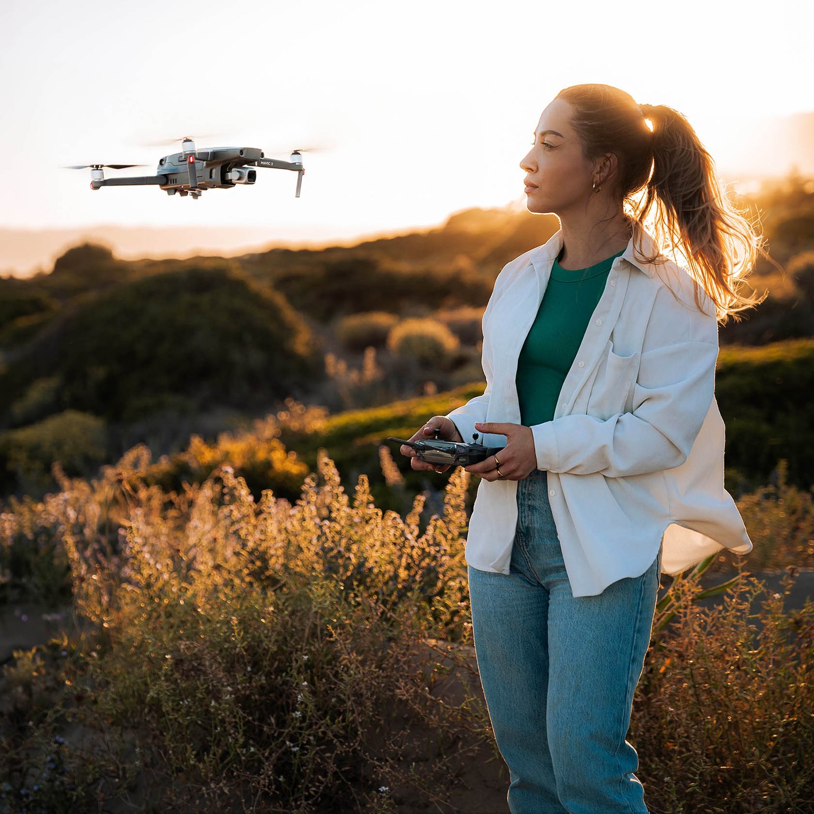 A woman stands in a sunlit outdoor setting with shrubs and hills in the background, representing Justina Kris Photography with a drone mid-flight with a remote in her hands. She is wearing a white shirt, green top, and jeans, with her hair in a ponytail.