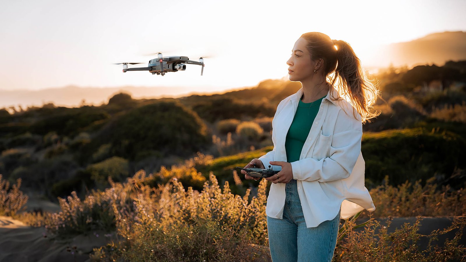 A woman with a ponytail, wearing a white shirt and jeans, operates a drone using a remote controller. The drone hovers in front of her at sunset, casting a warm glow over the scenic, bushy landscape. Justina Kris Photography captures this serene moment of technology blending with nature.