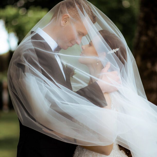 A couple stands closely, embracing each other tenderly under a sheer, flowing wedding veil that drapes over them. They gaze into each other's eyes, smiling softly. Lush green trees form a beautiful backdrop, adding to the romantic ambiance.