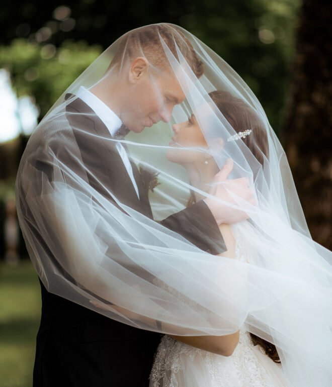 A couple stands closely, embracing each other tenderly under a sheer, flowing wedding veil that drapes over them. They gaze into each other's eyes, smiling softly. Lush green trees form a beautiful backdrop, adding to the romantic ambiance.