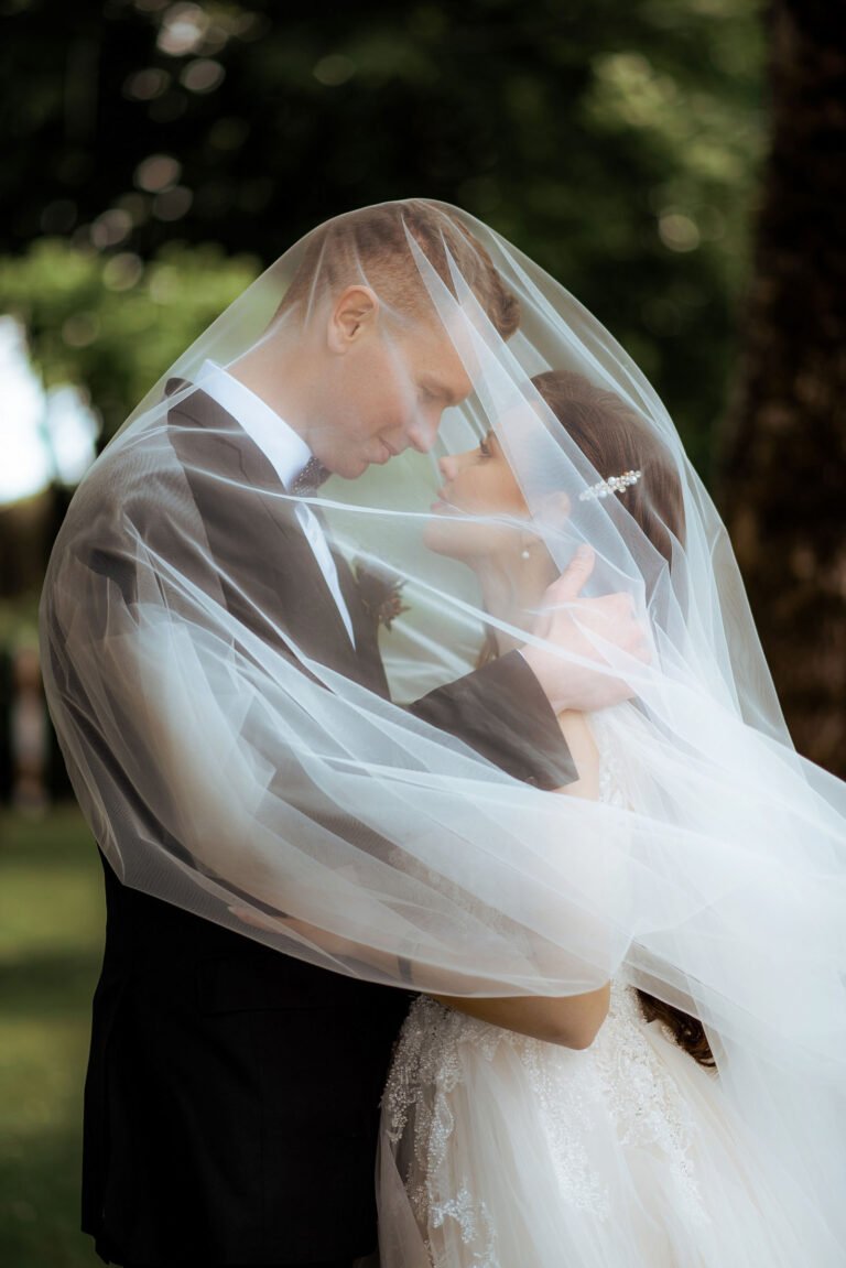 A couple stands closely, embracing each other tenderly under a sheer, flowing wedding veil that drapes over them. They gaze into each other's eyes, smiling softly. Lush green trees form a beautiful backdrop, adding to the romantic ambiance.