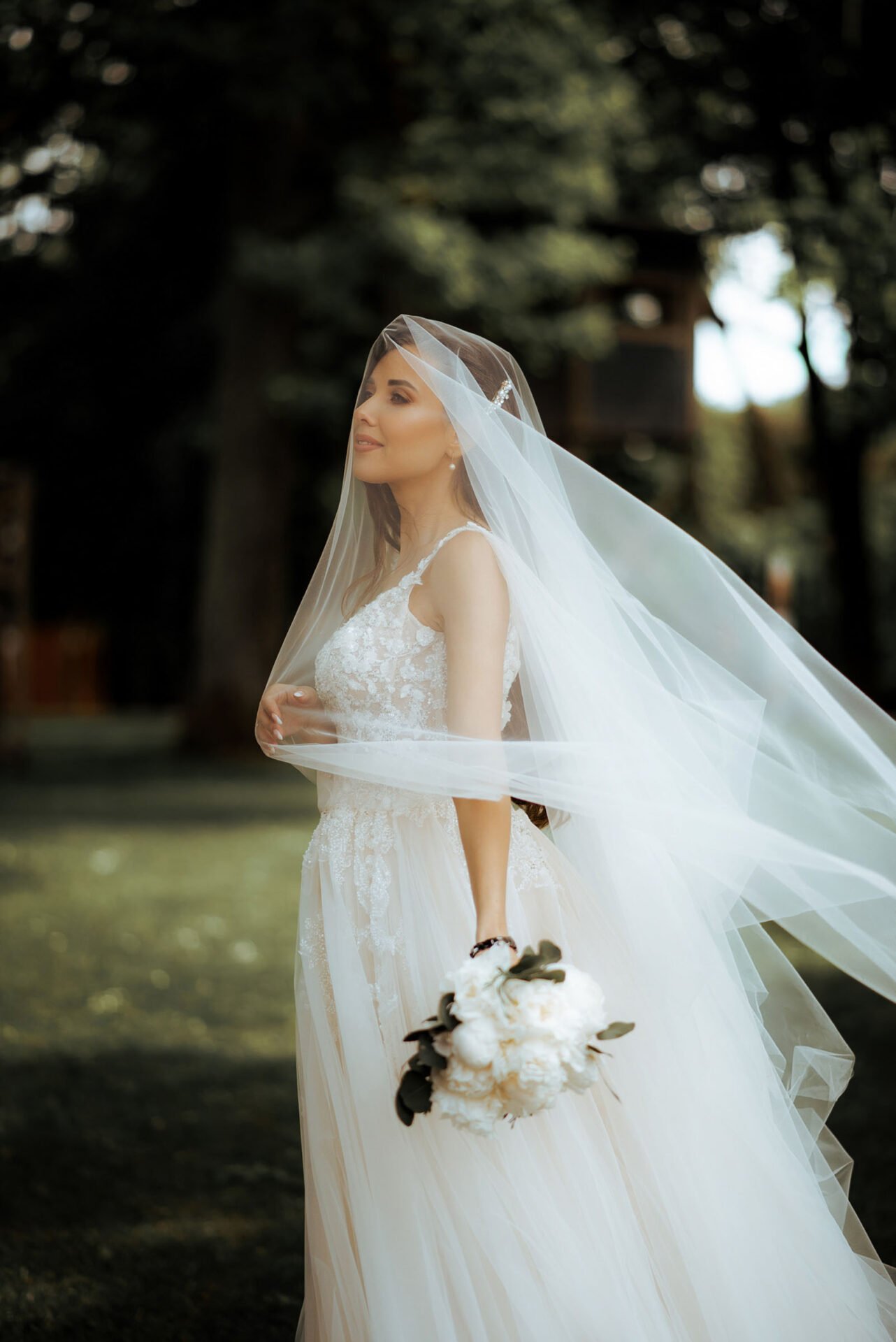 A bride stands outdoors in Marbella, wearing a white lace wedding dress and a long veil flowing in the breeze. She holds a bouquet of white flowers and gazes thoughtfully into the distance. The background is blurred with trees and greenery, captured beautifully by Wedding Photography Marbella.