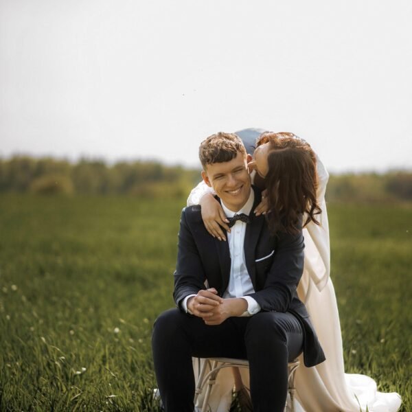 A person in a wedding dress hugs and kisses a person in a navy blue suit who is sitting on a chair in a grassy field. The person in the suit smiles broadly with their hands clasped in front of them. Captured by Luxury Wedding Photographer Marbella, the couple is surrounded by greenery with a blurred background.