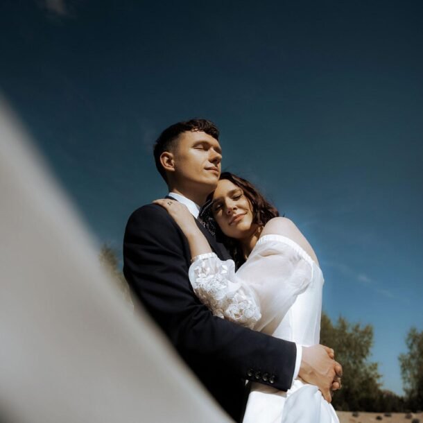 A couple dressed in wedding attire embraces outdoors under a clear blue sky. The bride, in a white gown with sheer sleeves, rests her head on the groom's chest, eyes closed. The groom, in a dark suit, gazes into the distance as trees stand tall behind them—a timeless moment captured by a wedding photographer from Costa del Sol.