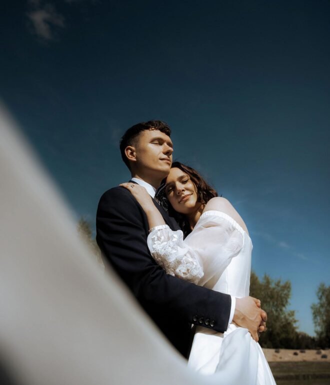 A couple dressed in wedding attire embraces outdoors under a clear blue sky. The bride, in a white gown with sheer sleeves, rests her head on the groom's chest, eyes closed. The groom, in a dark suit, gazes into the distance as trees stand tall behind them—a timeless moment captured by a wedding photographer from Costa del Sol.