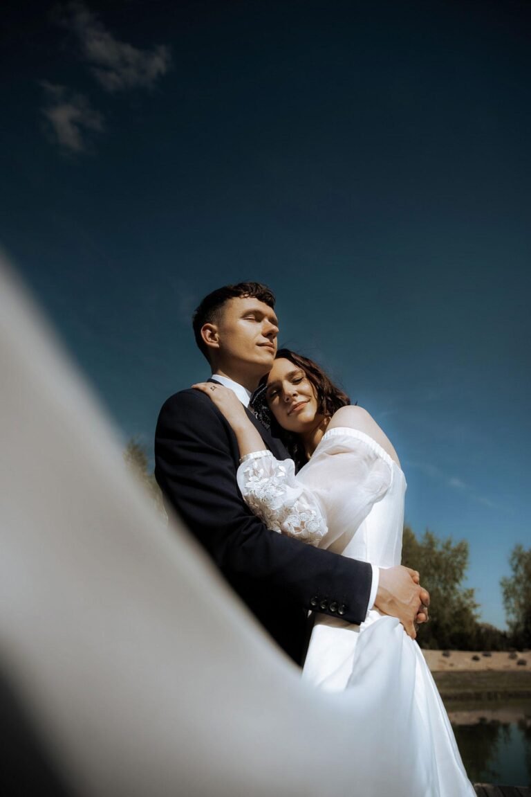 A couple dressed in wedding attire embraces outdoors under a clear blue sky. The bride, in a white gown with sheer sleeves, rests her head on the groom's chest, eyes closed. The groom, in a dark suit, gazes into the distance as trees stand tall behind them—a timeless moment captured by a wedding photographer from Costa del Sol.