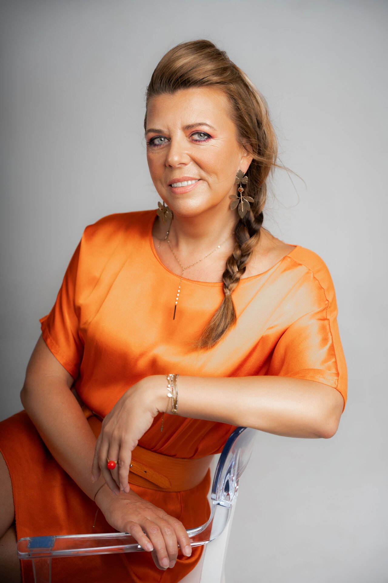 A woman with long, braided hair smiles at the camera. She is wearing a bright orange, short-sleeved dress and large, dangling earrings. Seated on a transparent chair with her left arm resting on the chair back, she displays bracelets on her wrist during a Branding Photoshoot Málaga session.