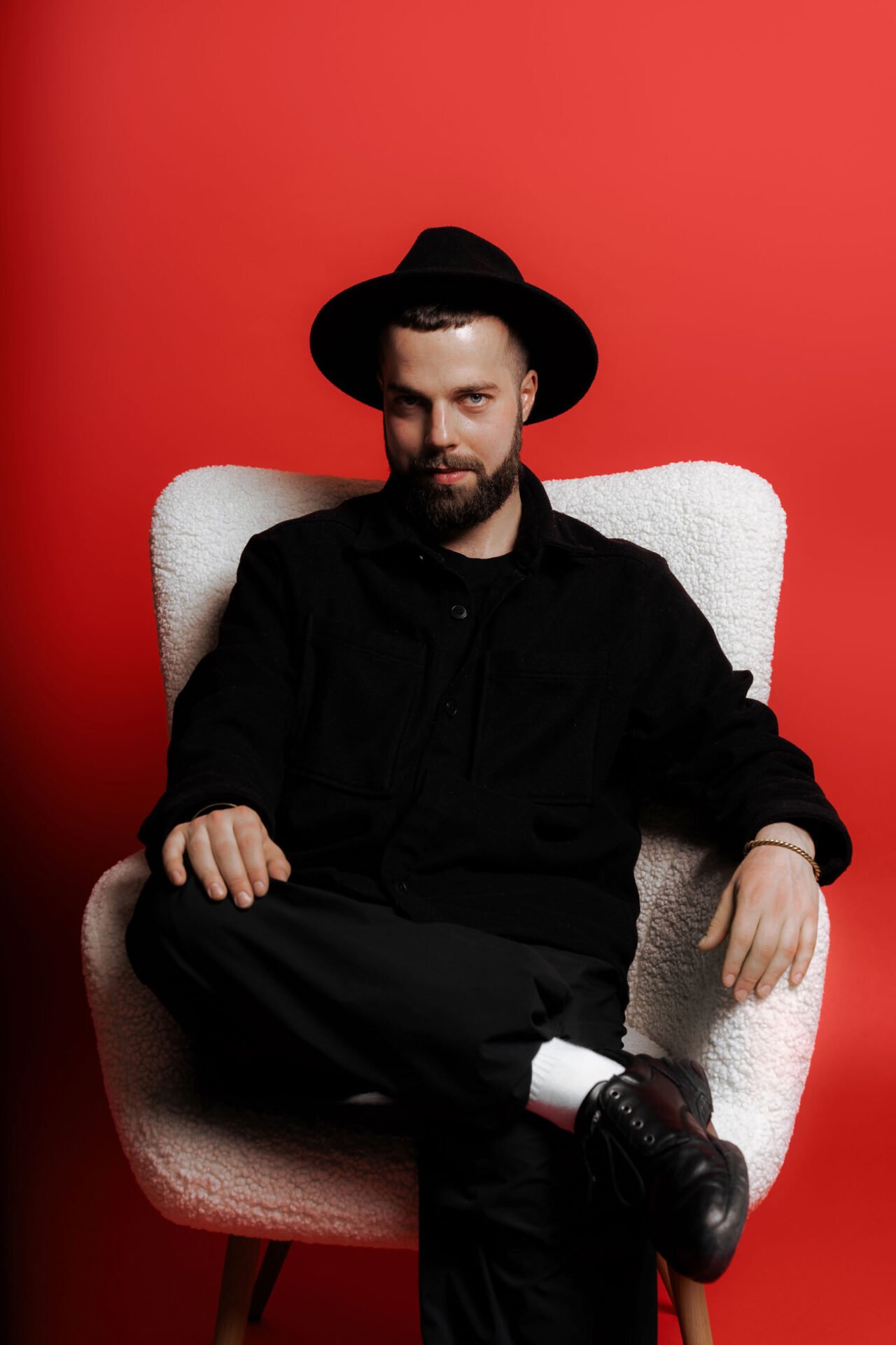 A man with a beard, wearing a black hat and black clothes, is sitting on a white textured armchair against a solid red background. He has one leg crossed over the other, exuding relaxed confidence during this business photoshoot.