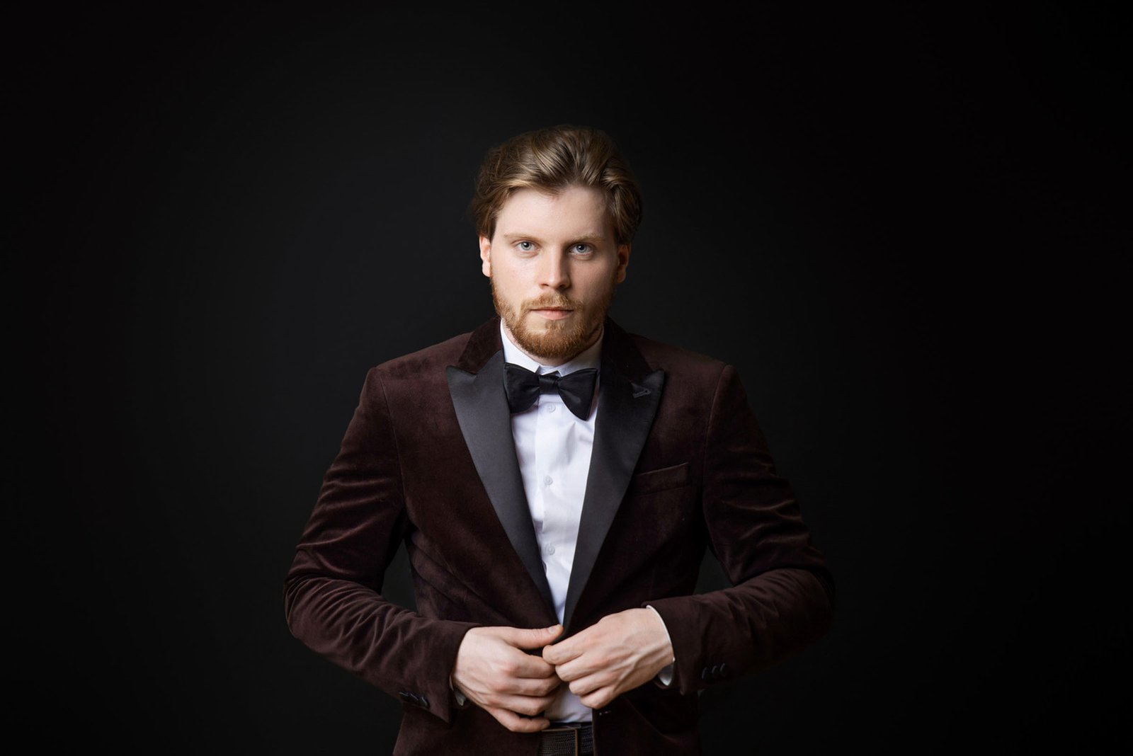 A man with a beard and styled hair stands against a dark background, wearing a dark velvet suit jacket, white dress shirt, and a black bow tie. He is buttoning his jacket and looking directly at the camera during a business photoshoot in Marbella.