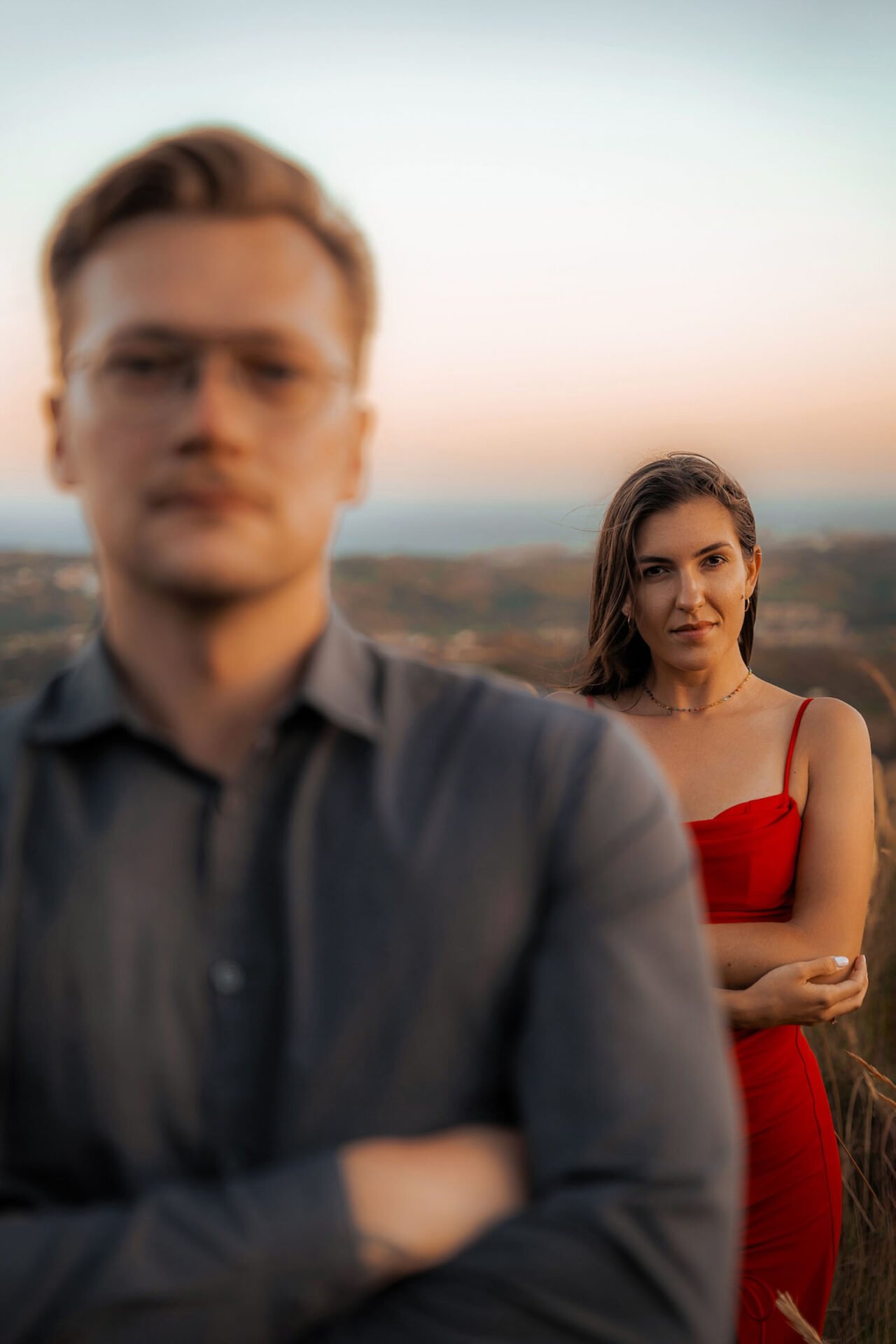 A man in glasses and a gray shirt stands with arms crossed in the foreground, out of focus. A woman in a red dress stands in the background, looking at the camera and slightly smiling. The setting appears to be an outdoor landscape with a soft sunset sky.