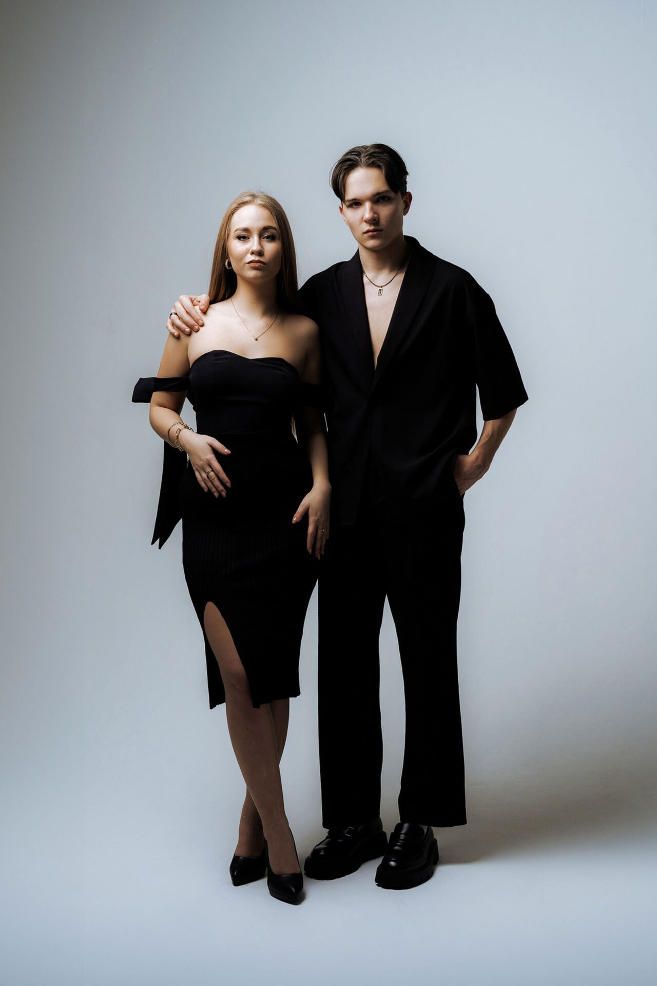 A woman and a man stand close together against a plain background during a photoshoot in Malaga. The woman is wearing a strapless black dress with a slit and black heels. The man is dressed in a loose black shirt, black trousers, and black shoes, with his arm around her shoulder.