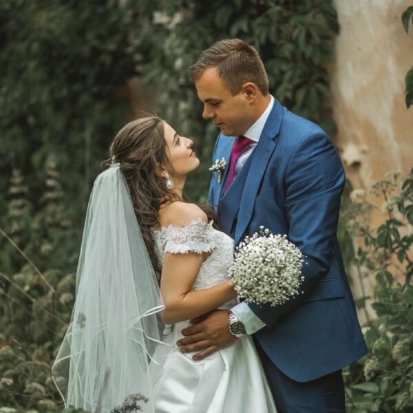 A bride and groom share an intimate moment outdoors in front of a wall covered in greenery. The bride, in an off-the-shoulder white gown and veil, holds a small bouquet of baby's breath. The groom, in a blue suit, lovingly embraces her with a tender look—captured flawlessly by a luxury wedding photographer Marbella.