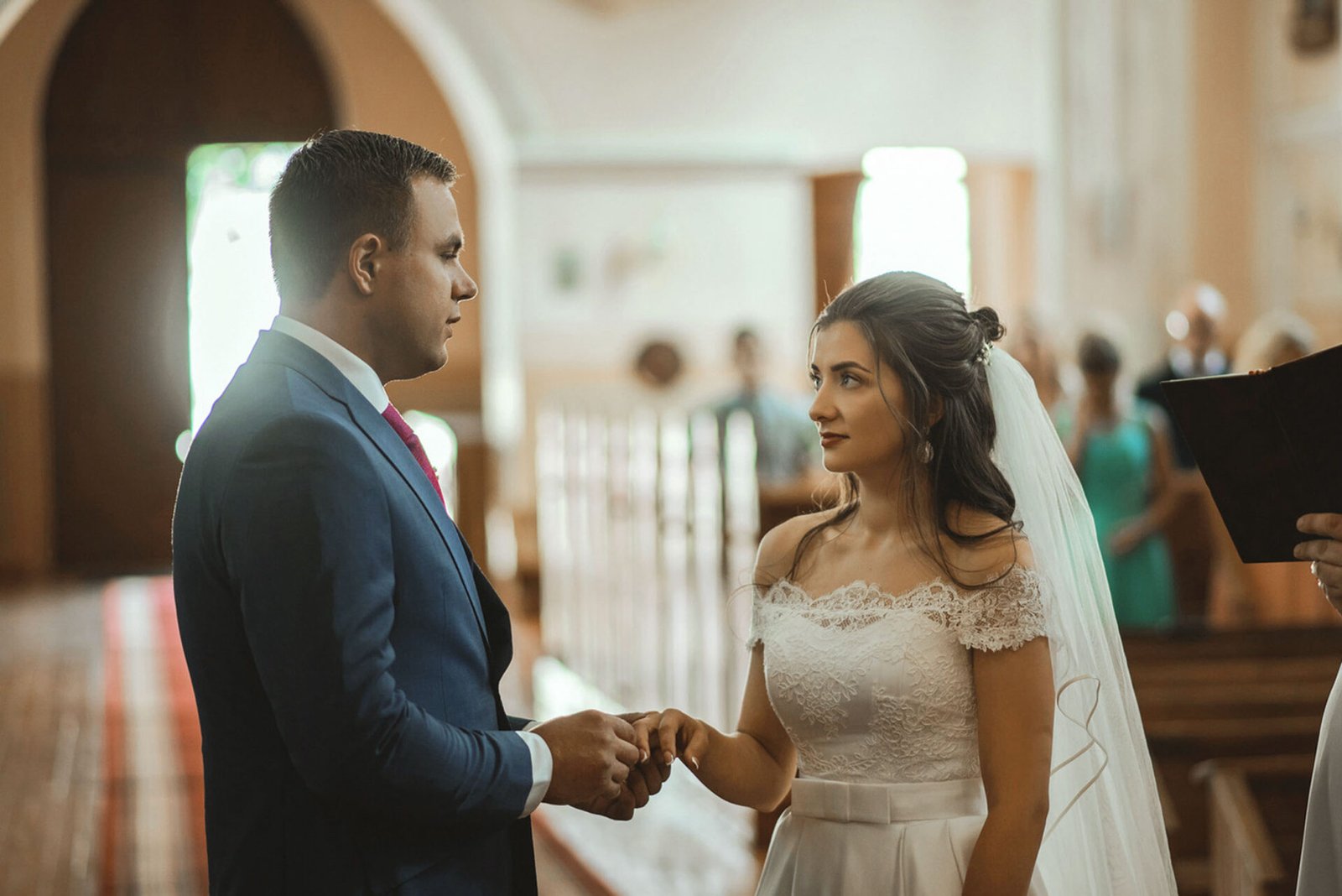 A bride, wearing a white dress with lace details and a veil, holds hands with the groom, dressed in a blue suit and red tie, during a wedding ceremony in a church. They look at each other intently, with guests blurred in the background—captured perfectly by a Wedding Photographer Costa del Sol.