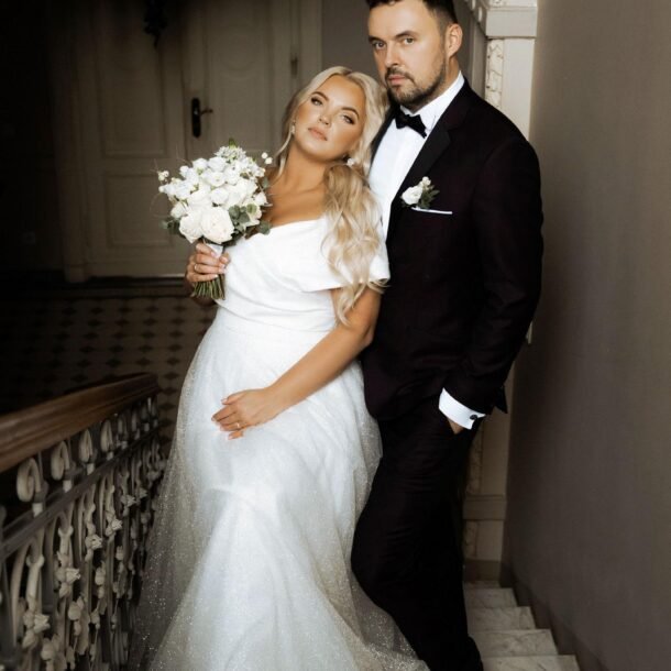 A bride and groom pose on an ornate staircase. The bride, wearing a white off-the-shoulder wedding gown and holding a bouquet of white flowers, leans against the groom. The groom, dressed in a dark suit with a white dress shirt, stands next to her with his hand in his pocket.