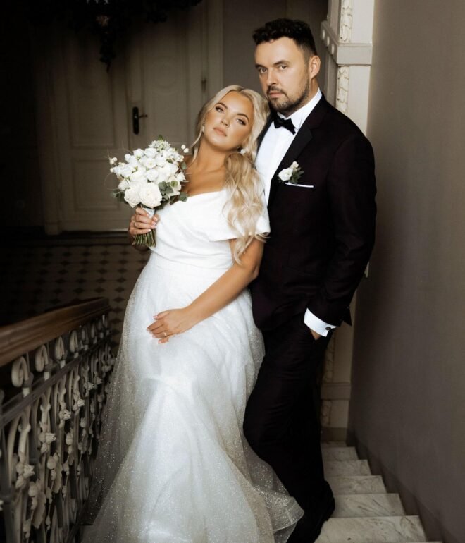 A bride and groom pose on an ornate staircase. The bride, wearing a white off-the-shoulder wedding gown and holding a bouquet of white flowers, leans against the groom. The groom, dressed in a dark suit with a white dress shirt, stands next to her with his hand in his pocket.