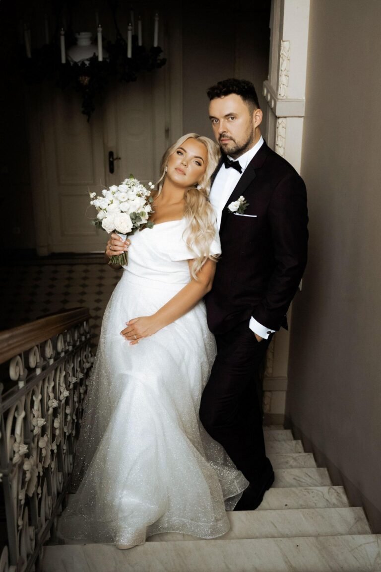 A bride and groom pose on an ornate staircase. The bride, wearing a white off-the-shoulder wedding gown and holding a bouquet of white flowers, leans against the groom. The groom, dressed in a dark suit with a white dress shirt, stands next to her with his hand in his pocket.