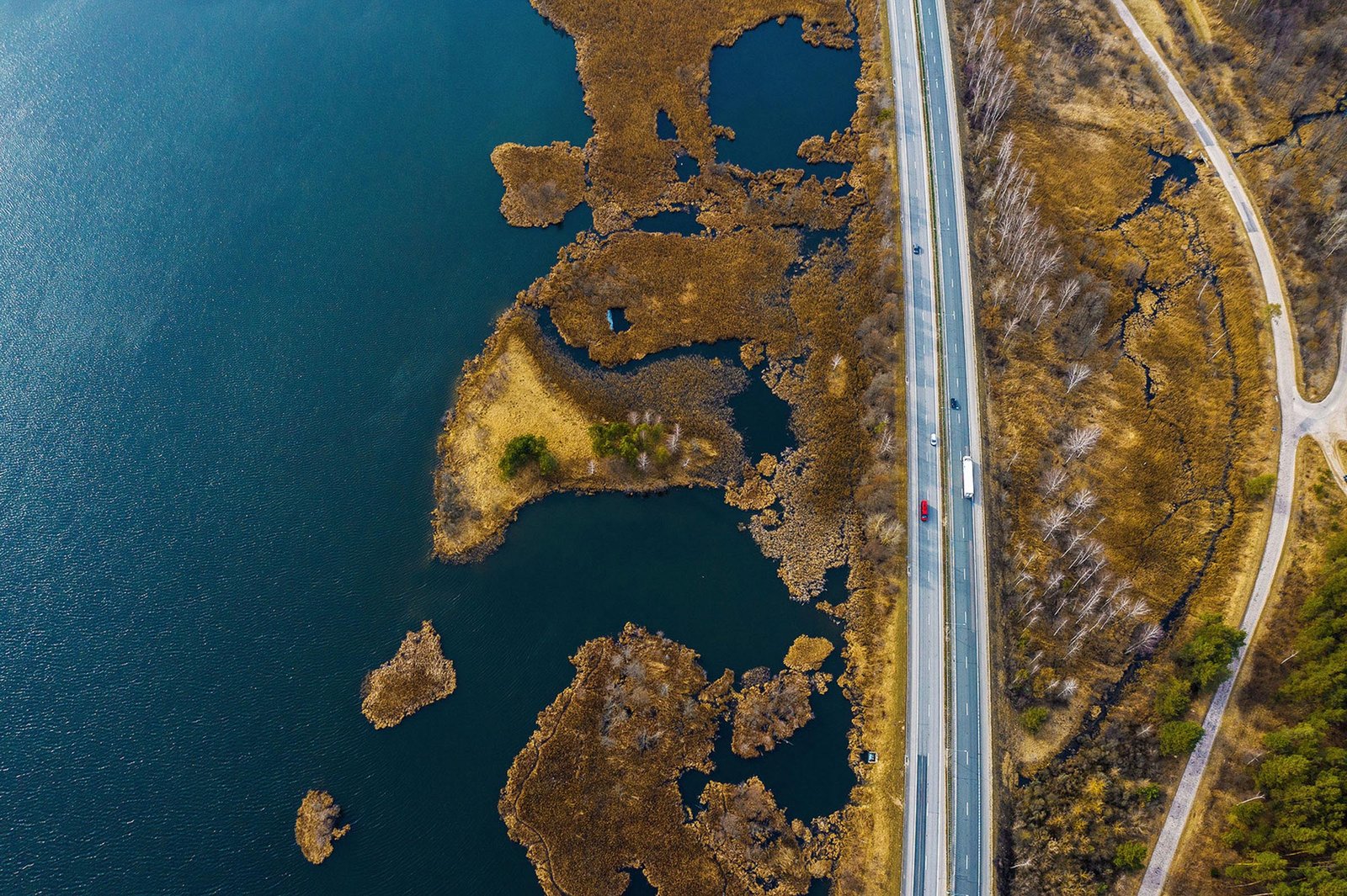 Aerial drone photography captures a blue lake with brown marshy islands. To the right, a straight road runs parallel to the lake, showcasing a red car and another vehicle. The landscape features dry vegetation and a mix of green trees.