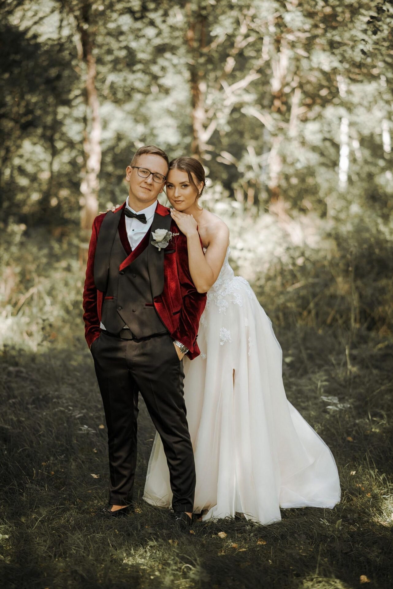 A bride in a white lace dress and a groom in a black suit with a red velvet jacket stand in a forested area. The groom has one hand in his pocket while the bride rests her head on his shoulder, wrapping her arm around his neck. They both have a serene expression.