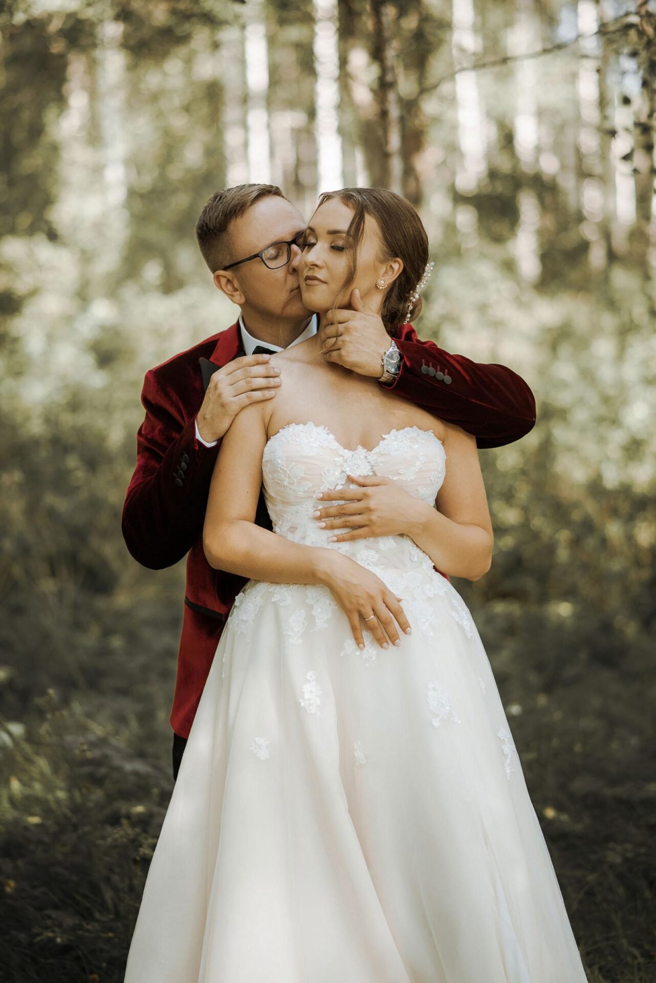 A bride and groom embrace in a forest. The groom, wearing a red velvet jacket and glasses, stands behind the bride, gently holding her chin and waist. The bride, in a lace strapless gown, holds his hand on her waist. Trees and sunlight create a serene backdrop—perfect for Wedding Photography Marbella.