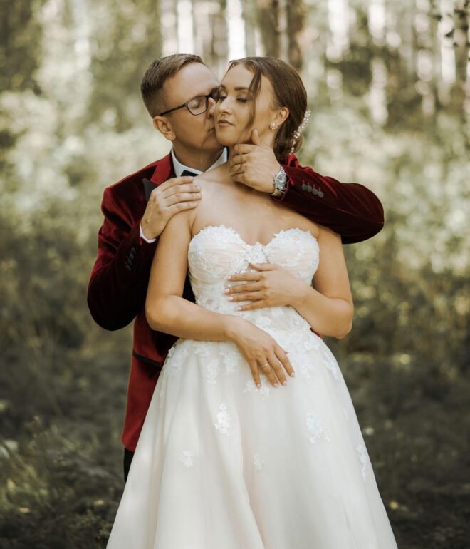 A bride and groom embrace in a forest. The groom, wearing a red velvet jacket and glasses, stands behind the bride, gently holding her chin and waist. The bride, in a lace strapless gown, holds his hand on her waist. Trees and sunlight create a serene backdrop—perfect for Wedding Photography Marbella.