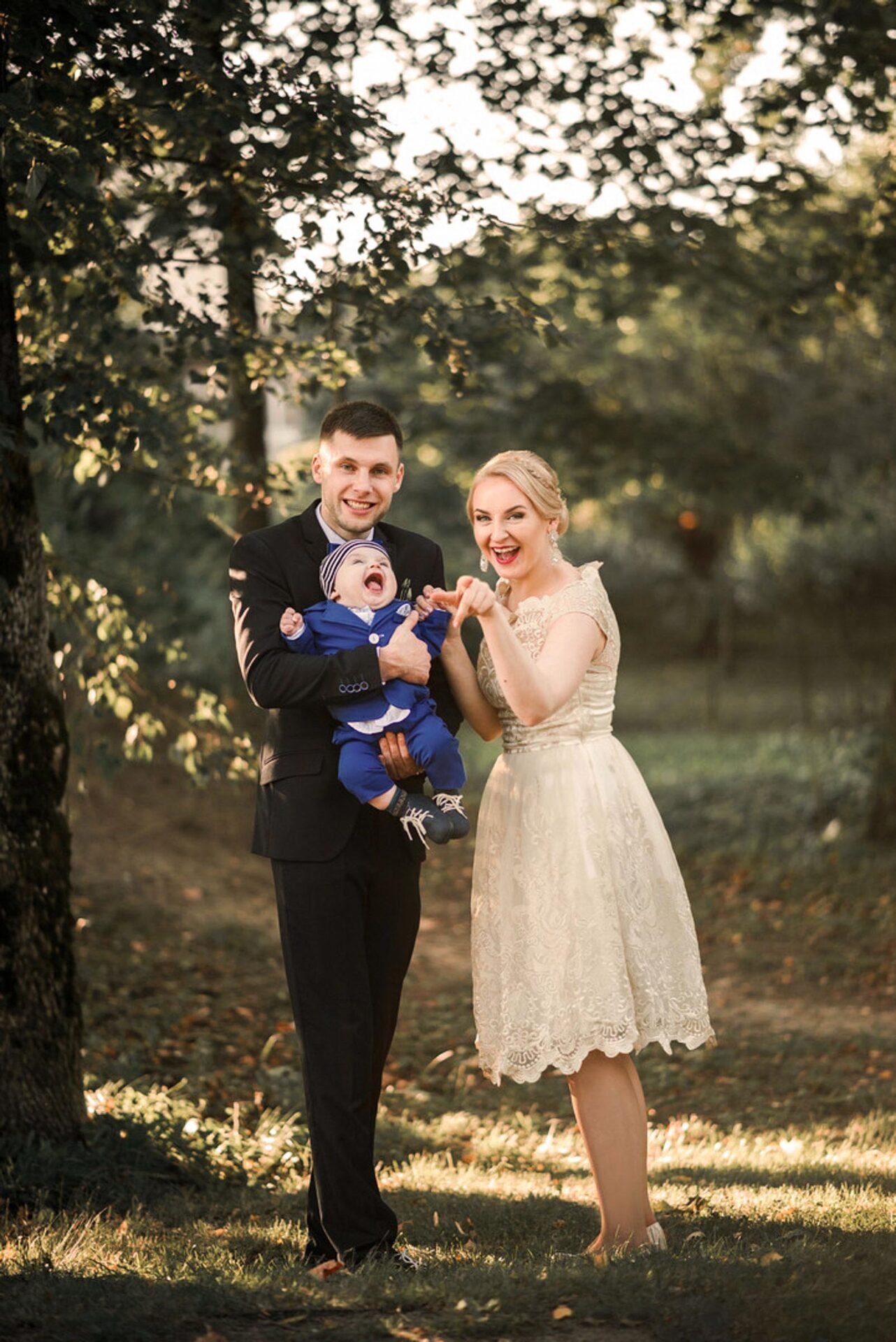 A family stands outdoors in a park-like setting, bathed in warm sunlight. The father, in a dark suit, holds a young child wearing a blue outfit and hat. The mother, in a cream-colored lace dress, stands nearby, both parents smiling joyfully at the camera.