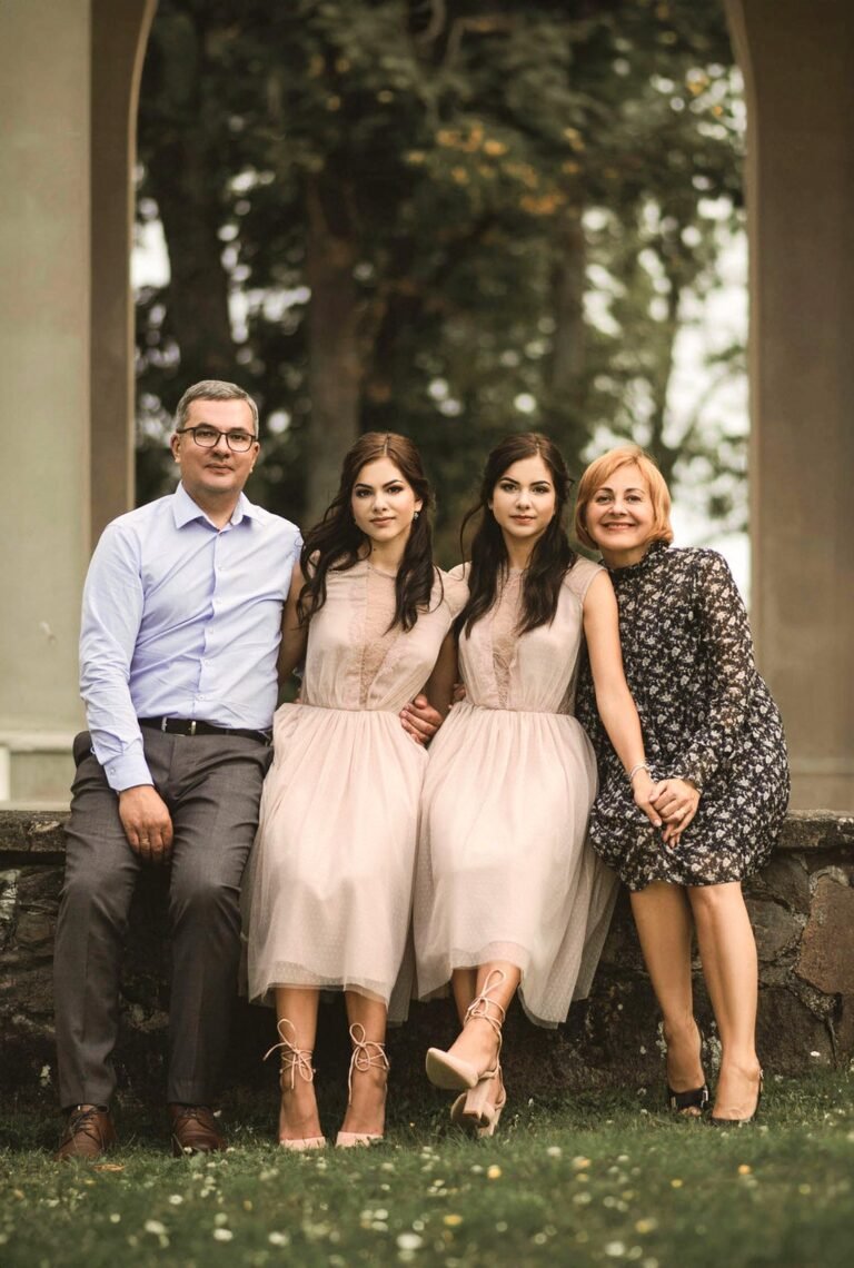 A group photo of four people sitting on a stone ledge outdoors. From left to right: a man in a blue shirt and dark pants, two women wearing matching pink dresses, and a woman in a patterned black dress. There are trees and an archway in the background.