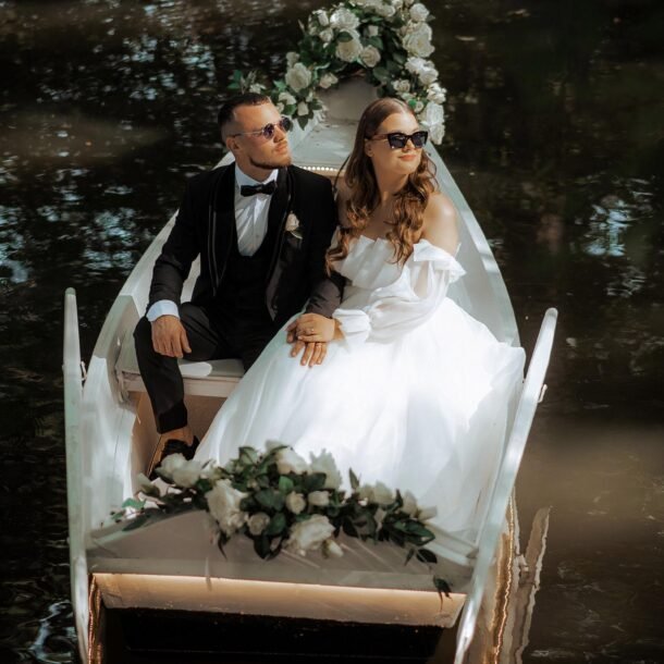 A bride and groom sit in a small, white boat decorated with flowers, floating on a gently rippling body of water. The bride is in a white dress and the groom in a black suit. Both wear sunglasses and gaze thoughtfully ahead. The scene is serene and romantic.