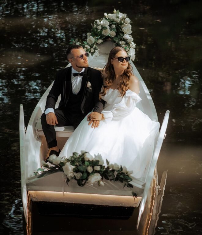 A bride and groom sit in a small, white boat decorated with flowers, floating on a gently rippling body of water. The bride is in a white dress and the groom in a black suit. Both wear sunglasses and gaze thoughtfully ahead. The scene is serene and romantic.