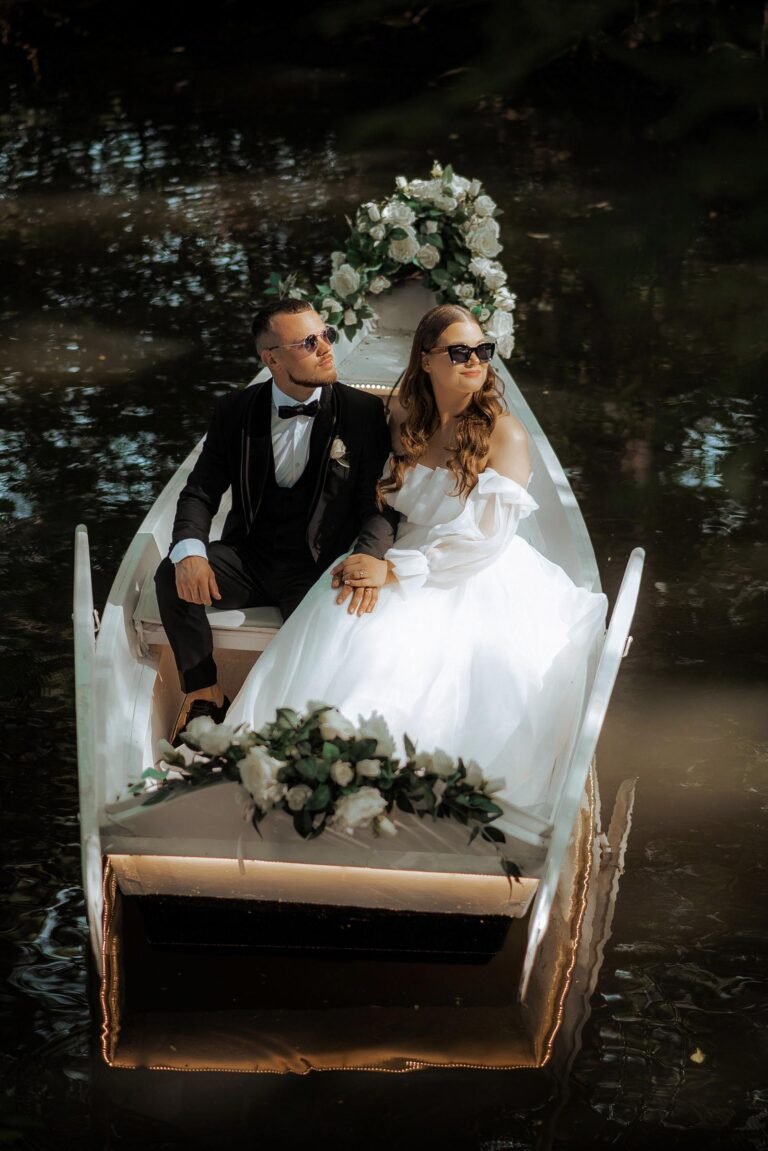 A bride and groom sit in a small, white boat decorated with flowers, floating on a gently rippling body of water. The bride is in a white dress and the groom in a black suit. Both wear sunglasses and gaze thoughtfully ahead. The scene is serene and romantic.