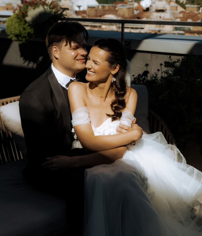 A couple wearing formal wedding attire sits closely on an outdoor sofa. The groom, in a black tuxedo, smiles at the bride, who is wearing an off-shoulder white gown. They are surrounded by lush greenery and the sun casts a warm glow, enhancing their joyful expressions – perfectly captured by a luxury wedding photographer in Marbella.