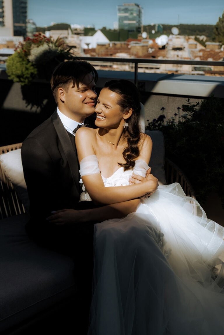 A couple wearing formal wedding attire sits closely on an outdoor sofa. The groom, in a black tuxedo, smiles at the bride, who is wearing an off-shoulder white gown. They are surrounded by lush greenery and the sun casts a warm glow, enhancing their joyful expressions – perfectly captured by a luxury wedding photographer in Marbella.