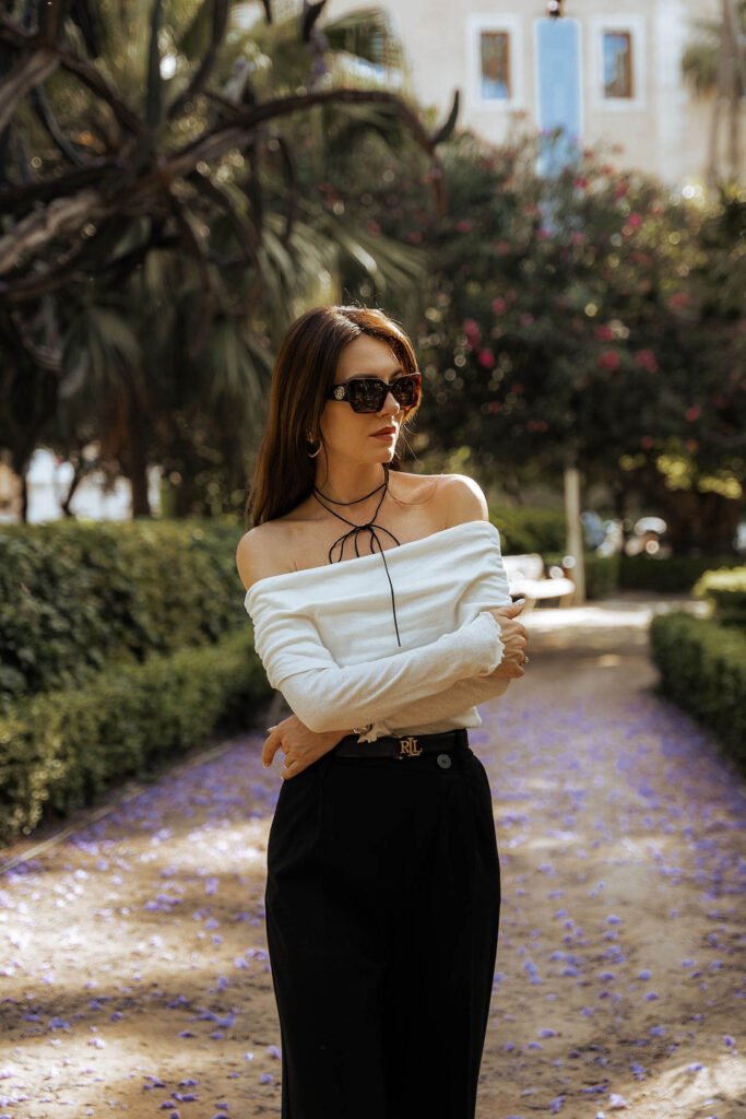 Woman in a white off-the-shoulder top and black pants, standing on a path lined with purple flowers, showcasing spring fashion in the city park of Malaga.