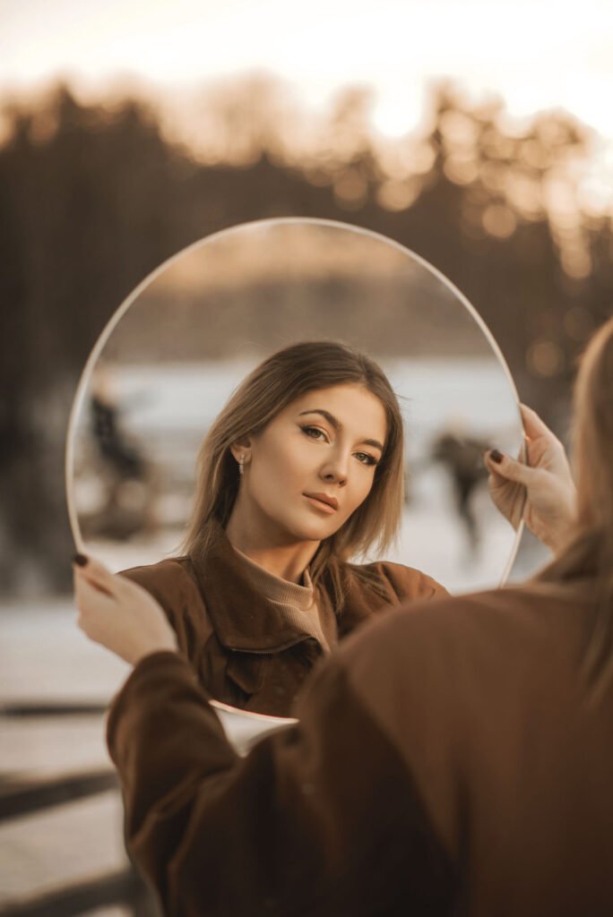 A woman with long hair looks into a circular mirror she holds up with both hands. The scene is outdoors during sunset, with blurred trees and people in the background. She wears a brown coat and has a serene expression.