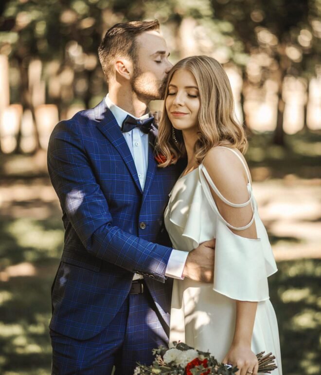 A couple stands outside, with the man in a blue plaid suit gently kissing the woman on the forehead. The woman, in a white, off-the-shoulder dress, holds a colorful bouquet. They are surrounded by greenery, creating a serene, romantic atmosphere.