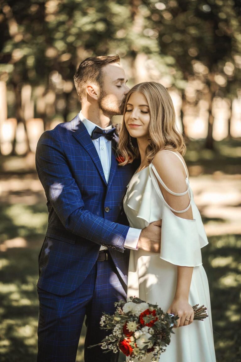 A couple stands outside, with the man in a blue plaid suit gently kissing the woman on the forehead. The woman, in a white, off-the-shoulder dress, holds a colorful bouquet. They are surrounded by greenery, creating a serene, romantic atmosphere.