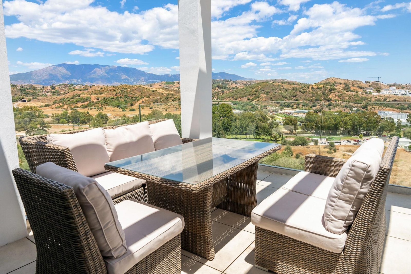 Spacious outdoor terrace with a wicker dining set featuring cushioned chairs and a glass-top table. Beyond the terrace is a scenic view of rolling hills, green trees, and mountain ranges under a partly cloudy blue sky, ideal for Real Estate Photography Marbella.