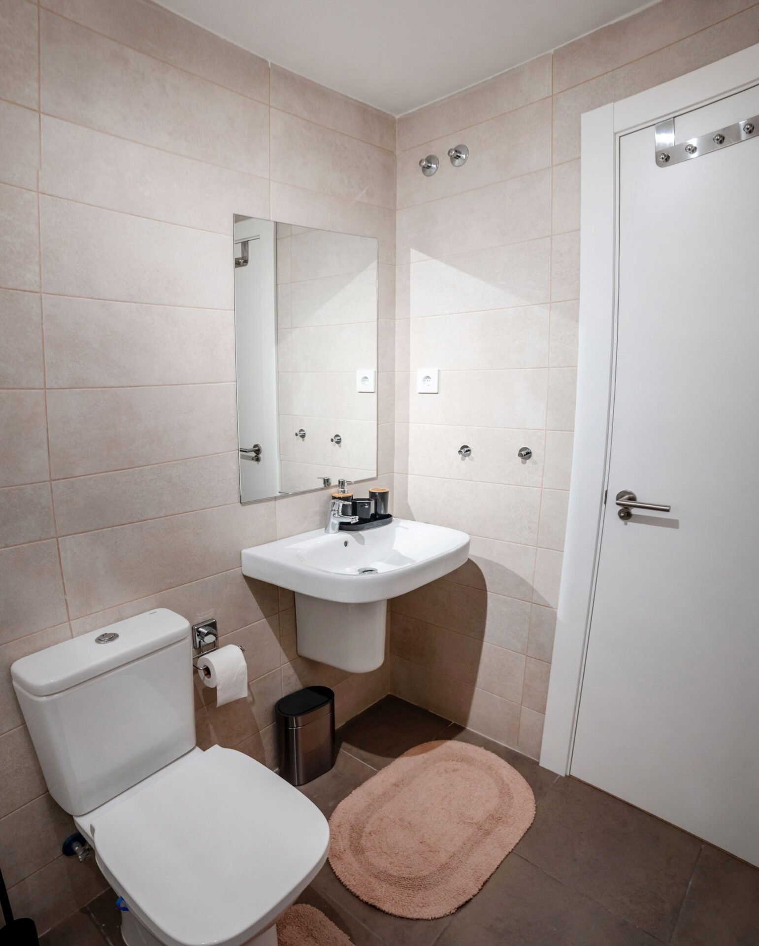 A small, modern bathroom with beige tile walls featuring a toilet, a sink with a mirror above it, and a white door. The floor has matching beige rugs in front of the toilet and sink. There are small chrome fixtures, including a toilet paper holder and a small trash bin.