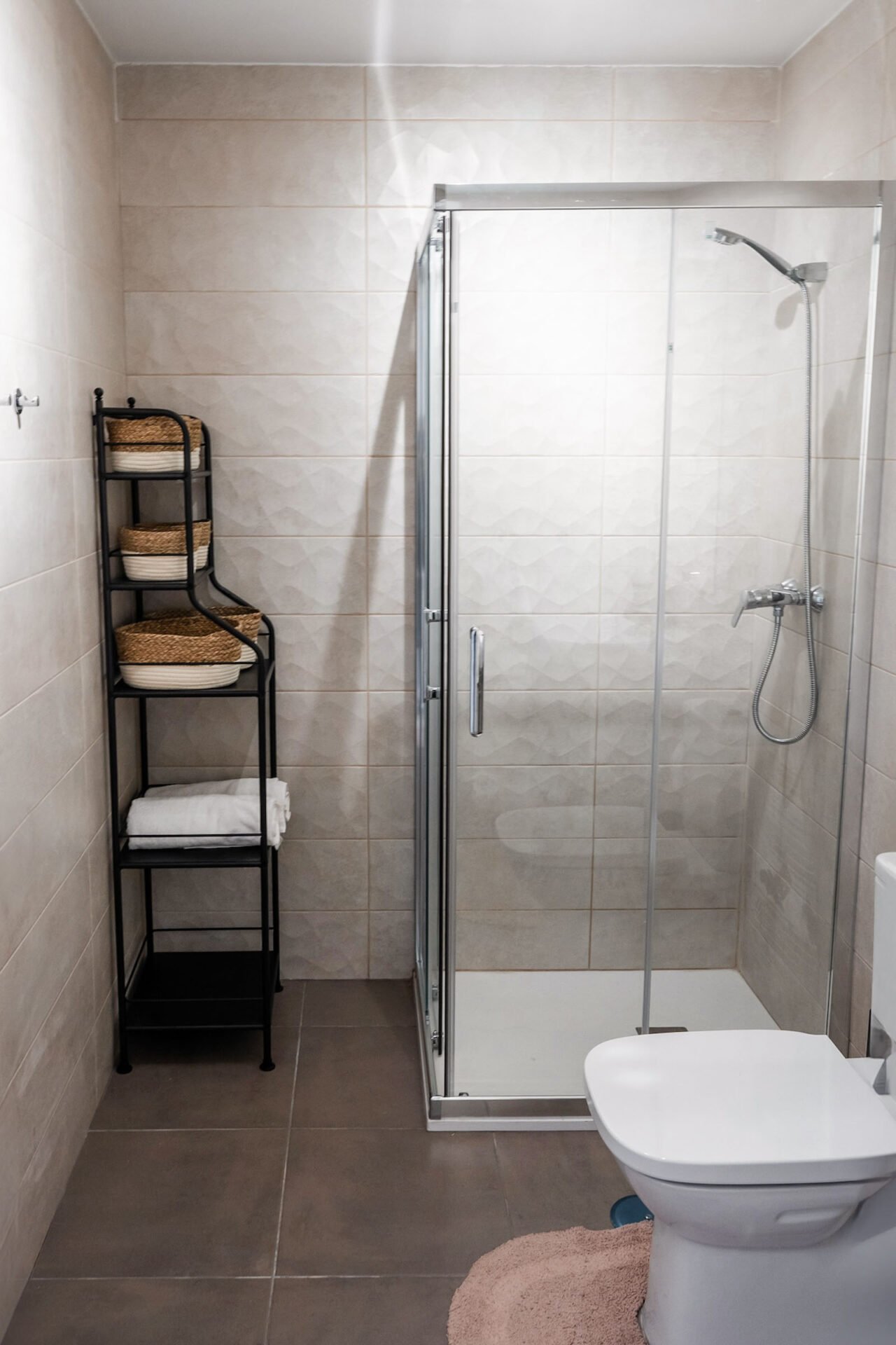 A modern bathroom featuring a glass shower enclosure with a chrome showerhead, a white toilet with a pink mat in front, and a black metal shelf unit holding wicker baskets and white towels against a tiled wall. With beige and gray hues, this space exemplifies the elegance captured in Interior Property Photography Costa del Sol.