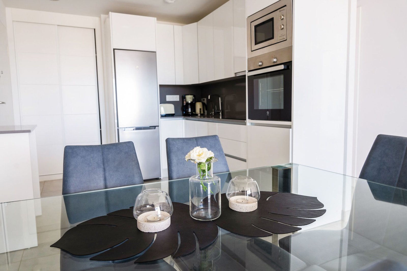 Modern kitchen with a glass dining table featuring a floral centerpiece and two candle holders on black placemats. The background shows white cabinetry, stainless steel appliances including a refrigerator, microwave, and an oven, and a coffee maker on the counter. Ideal for Real Estate Photography Marbella.