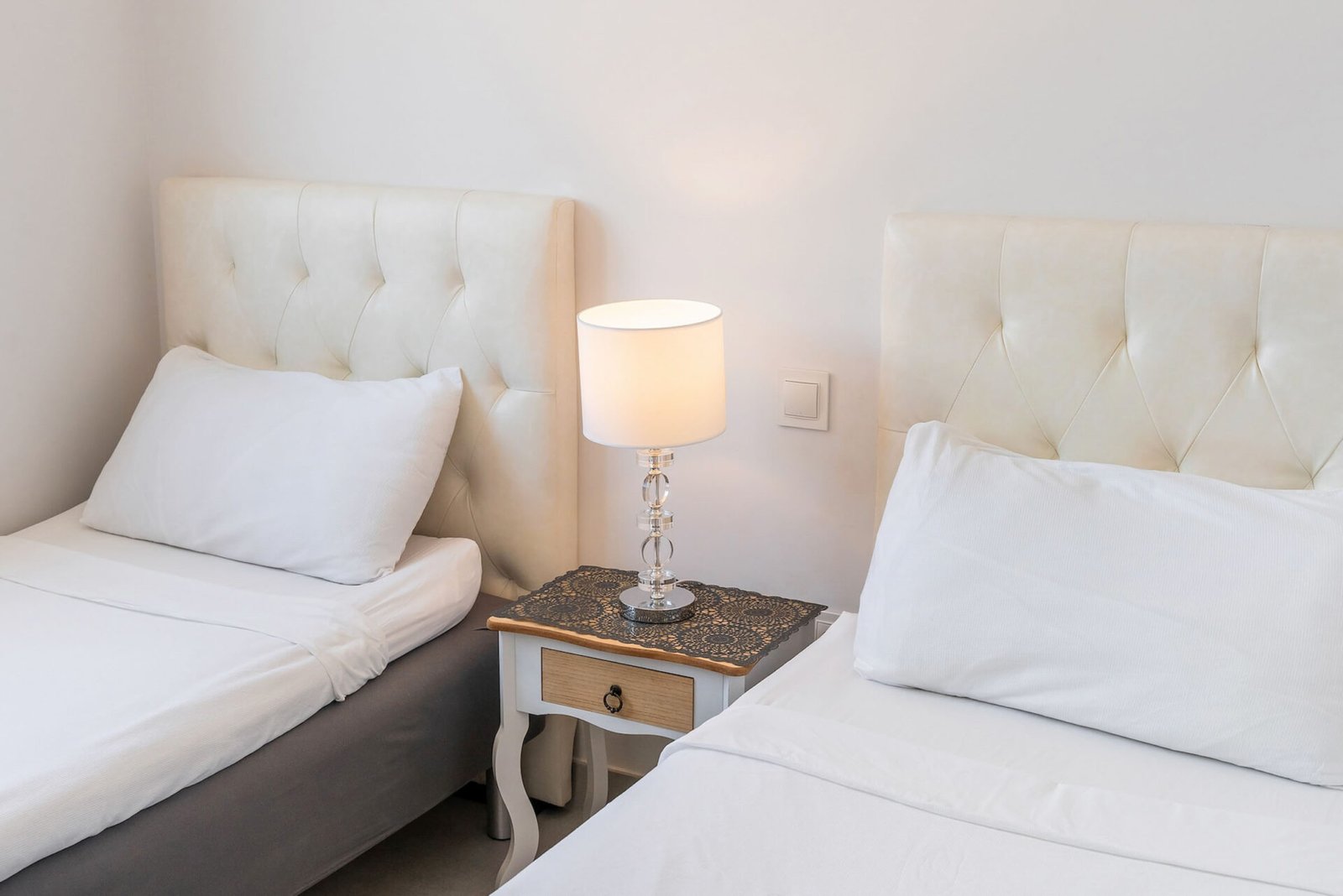 A minimalist bedroom with two single beds, each featuring a cushioned headboard and white bedding. Between the beds, a small nightstand with a drawer and an ornate tablecloth holds a decorative lamp with a white shade. The plain white walls offer an ideal setting for Real Estate Photography Marbella.