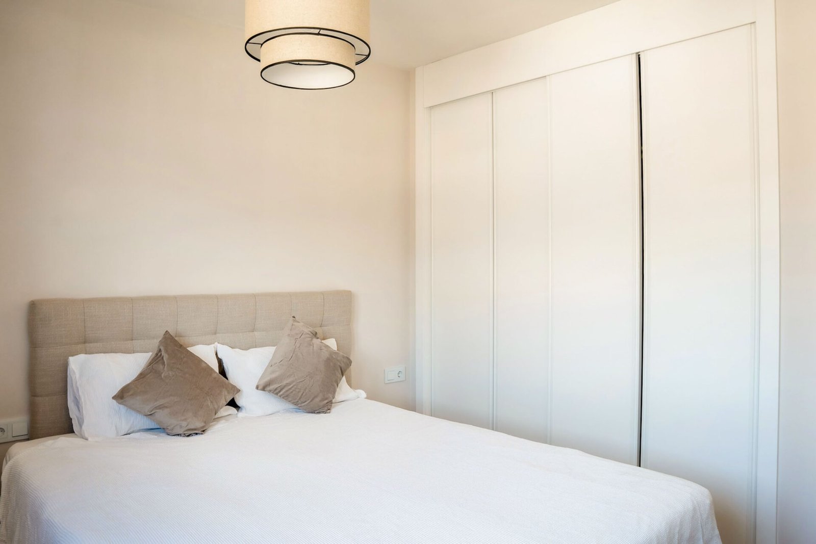 A minimalist bedroom features a neatly made bed with a beige upholstered headboard and two grey pillows resting against white pillows. The room, ideal for Interior Property Photography Costa del Sol, has white walls, a simple pendant light, and a closet with sliding white doors. The overall decor is clean and modern.