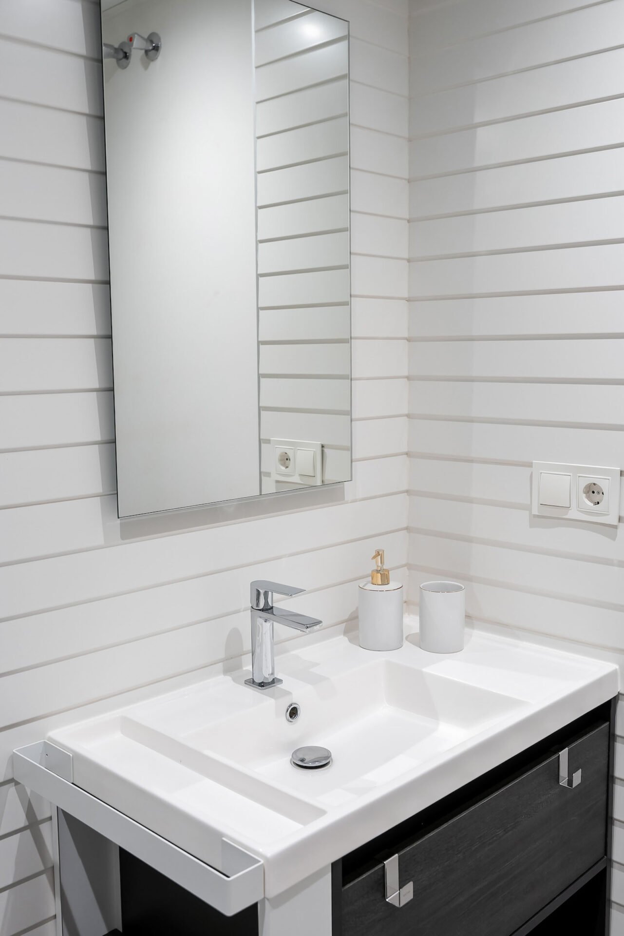 A modern bathroom sink with a rectangular mirror above it, perfect for Real Estate Photography in Marbella. The sink countertop holds a soap dispenser and a toothbrush holder. The walls feature white horizontal paneling, and there are two electrical outlets on the right side of the wall.