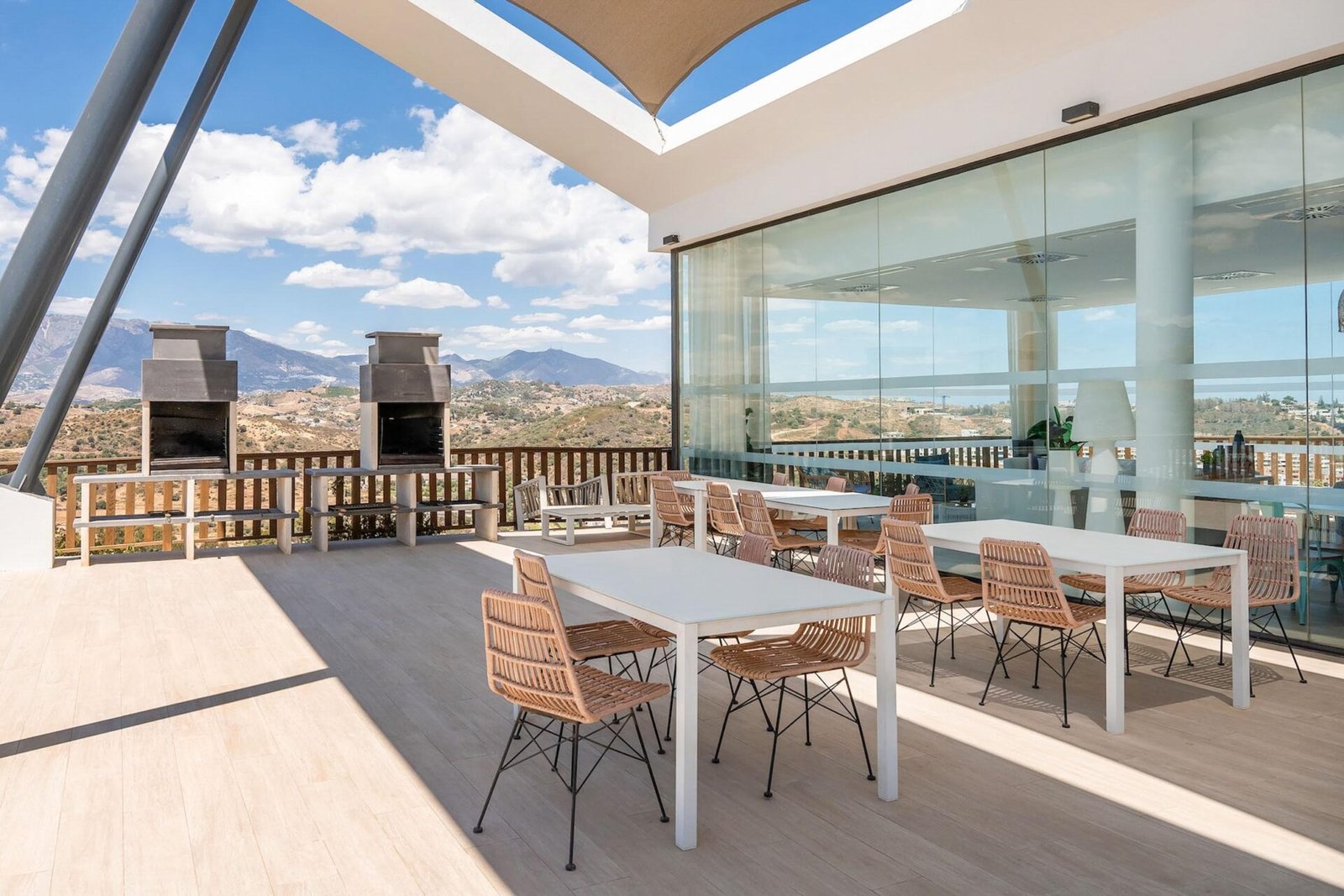 A stunning modern outdoor terrace with wicker chairs and white tables, set against a scenic mountain backdrop. The terrace features two large barbecue grills and is partially covered by a canopy. Glass walls reflect the clear blue sky and the surrounding landscape—perfect for Property Photography Costa del Sol.