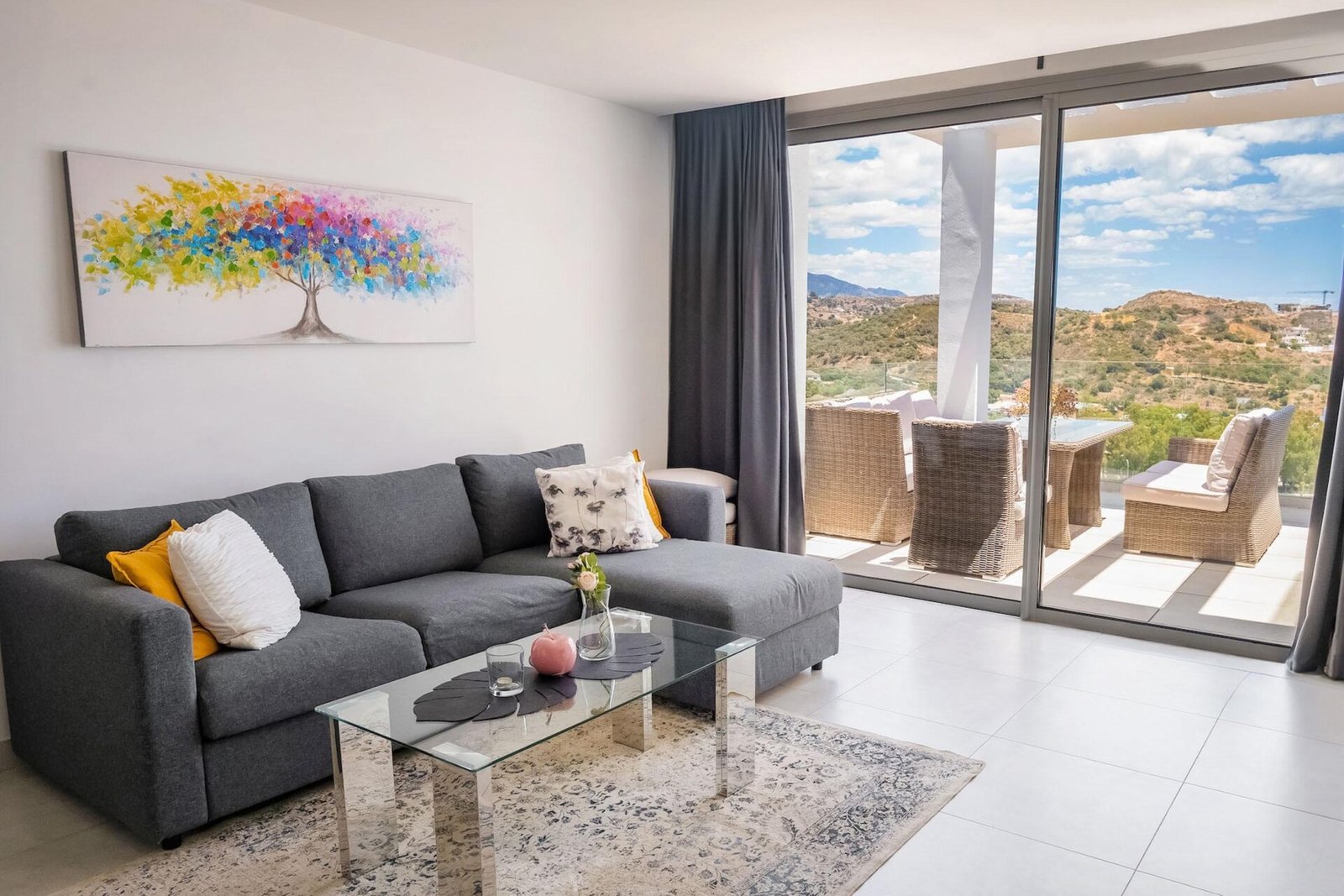 Modern living room with a gray sectional sofa adorned with colorful and white pillows. A glass coffee table is in front, and a vibrant abstract tree painting decorates the wall. Floor-to-ceiling windows open to a balcony with outdoor seating and a scenic view—perfect for Real Estate Photography Marbella.