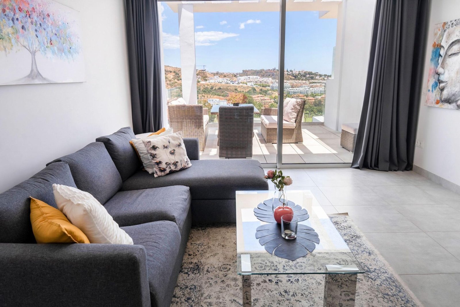 A modern living room featuring a grey sectional sofa adorned with colorful cushions, a glass coffee table with a red vase, and wall art. Large sliding glass doors open to a spacious balcony with outdoor seating, offering a scenic view of a hilly landscape—perfect for real estate photography in Marbella.