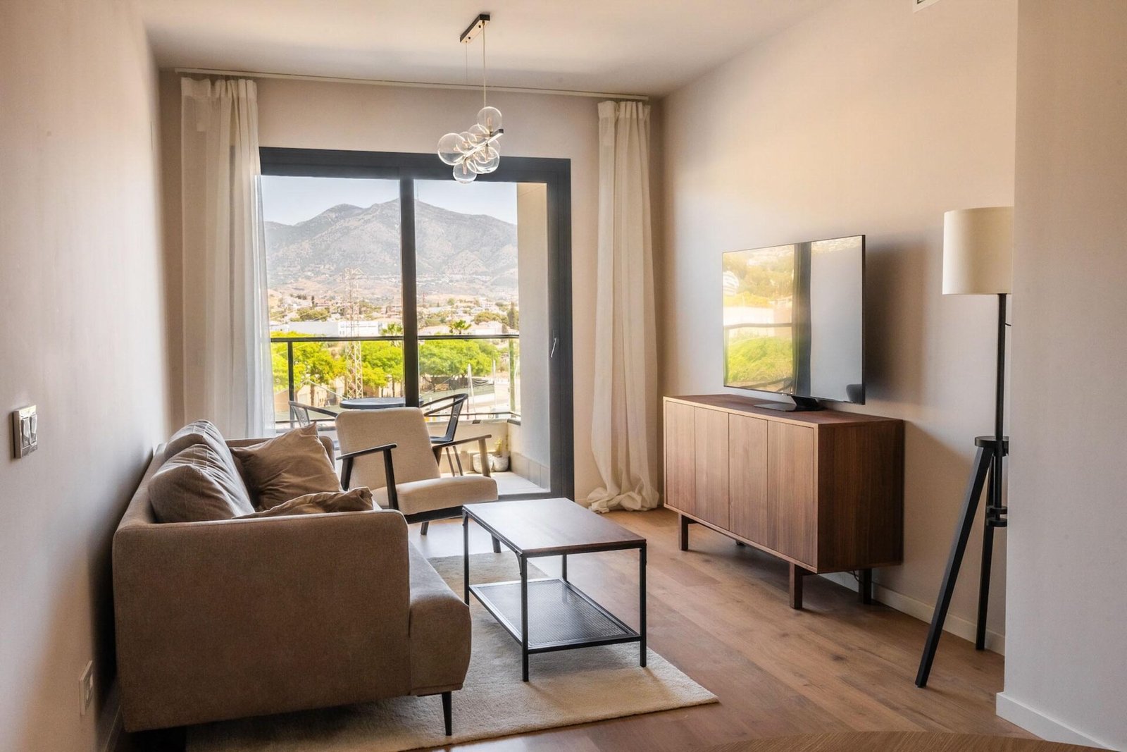 A cozy, modern living room with a beige sofa, a wooden coffee table, and a flat-screen TV on a wooden stand. A floor lamp stands beside the TV. Large sliding glass doors open to a balcony with a scenic mountain view. Natural light fills the room, perfect for Interior Property Photography Costa del Sol.