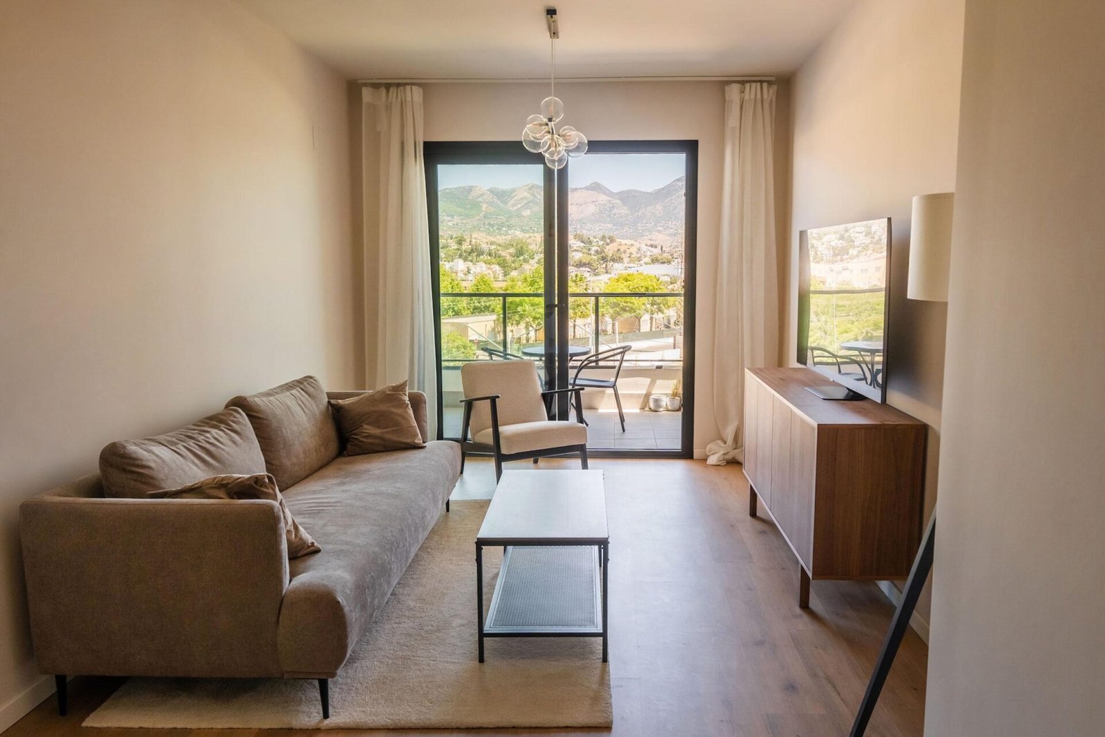 A modern living room featuring a gray sofa adorned with pillows, a glass coffee table, a cushioned chair, a TV mounted on a wooden console, and a black floor lamp. The room opens to a balcony with scenic mountain views and includes two patio chairs and a table—ideal for property photography Costa del Sol.