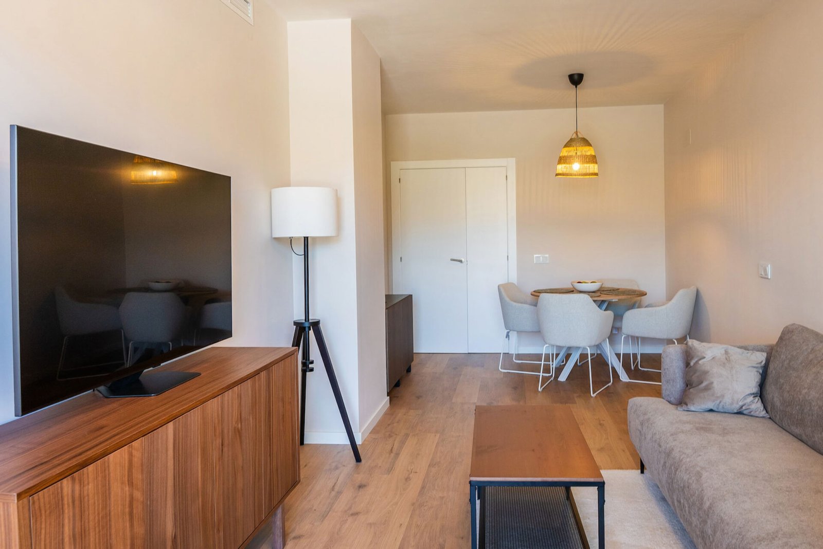 A modern living and dining area featuring a wall-mounted flat-screen TV on a wooden cabinet, a tall floor lamp, a gray sofa, and a wooden coffee table. The dining space includes a round table with four gray chairs under a woven pendant light, and a door in the background; perfect for real estate photography Marbella.