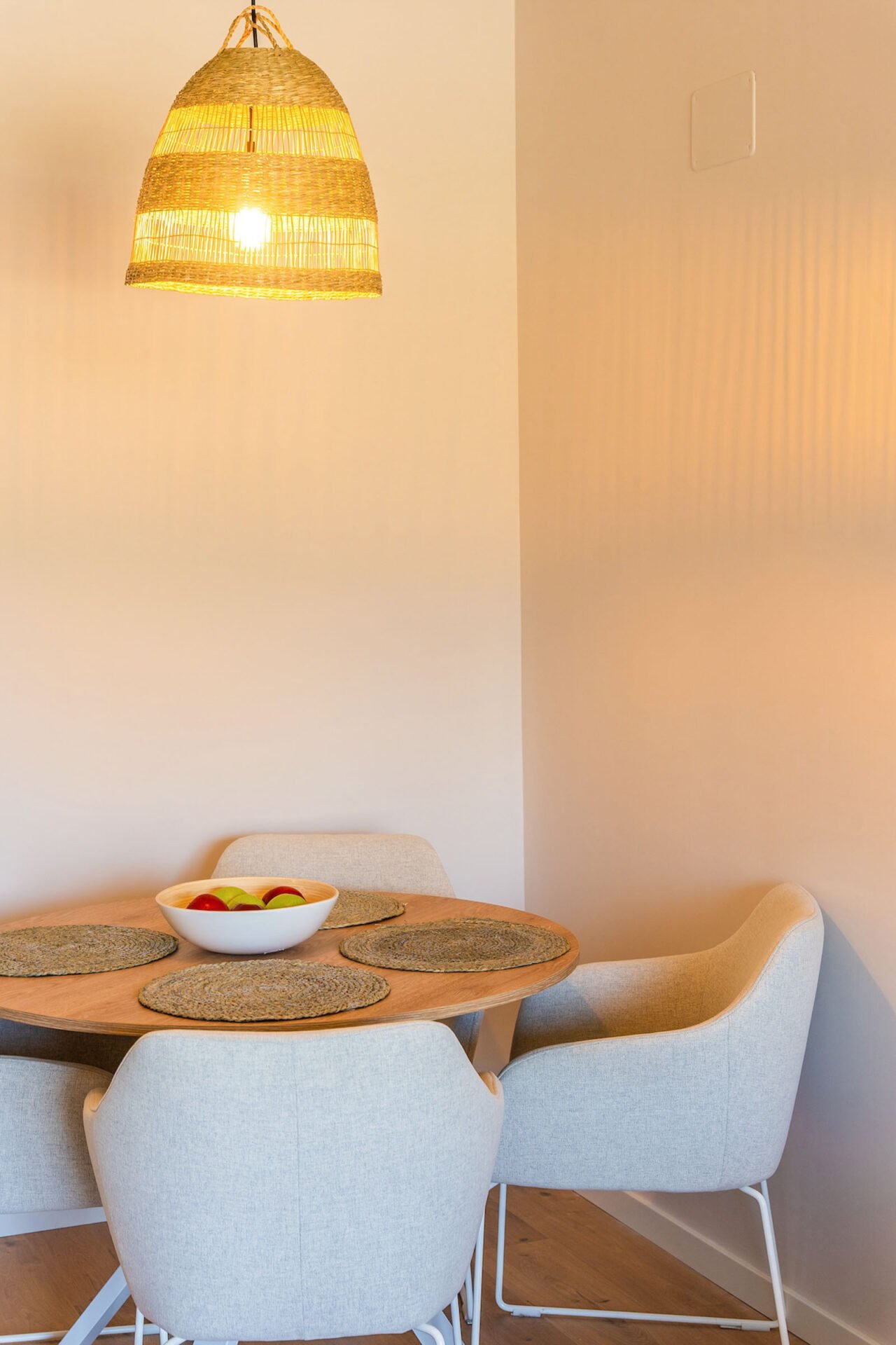 A cozy dining area with a round wooden table, four beige cushioned chairs, and a bowl of fruit on the table. A woven rattan pendant light hangs above, casting a warm glow. The white walls enhance the clean and inviting atmosphere—ideal for Interior Property Photography Costa del Sol.