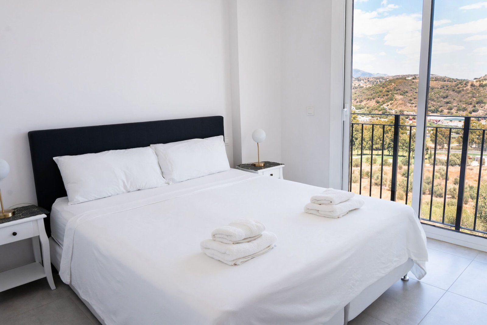 A bright bedroom with large windows overlooking a scenic landscape. The room features a neatly made bed with white linens and two stacks of folded towels at the foot. Small nightstands with lamps sit on either side of the bed, enhancing the minimalist decor—perfect for Real Estate Photography Marbella.